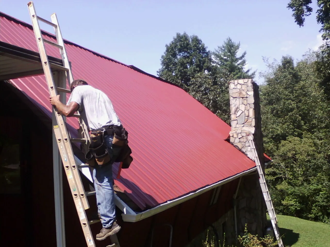Roofer on a ladder beside a red metal roof and stone chimney — Soffit & Fascia Repair by Roofing Brothers in Sparland